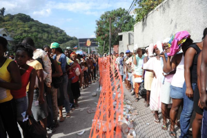 Lange Schlange von Menschen, die an einer Wahllokal-Schlange stehen, mit einem Zaun, einer Wand, Bäumen, Gebäuden, Strommasten, Drähten und einer Werbetafel im Hintergrund.
