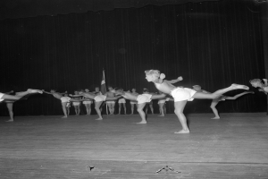 Schwarzes Foto einer Gruppe, die auf einer Bühne mit Vorhang und Flaggenhintergrund gymnastikübungen durchführt.
