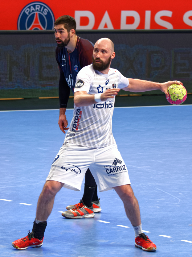 Zwei Männer spielen Handball auf einem Court, einer hält den Ball, mit einer Tafel im Hintergrund, die "Paris Saint-Germain vs Paris Saint Germain" anzeigt.