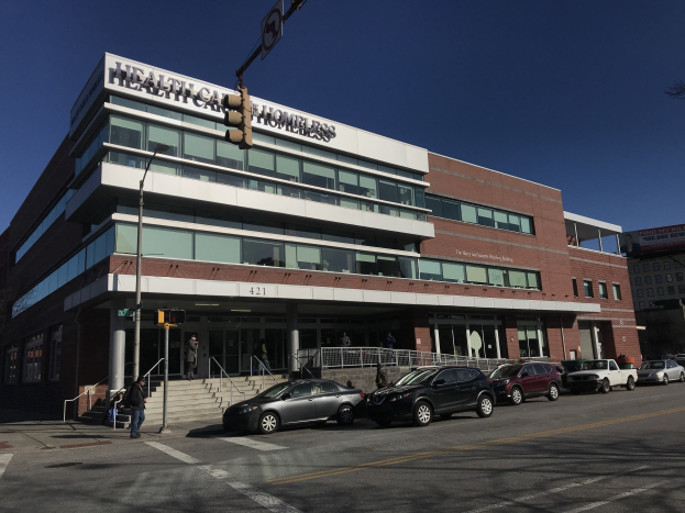 Gro├če Geb├Ąude mit dem Namen New York City Health Care Homeless Center, vor dem Autos geparkt sind, Menschen, Stra├čenm├Âbel und B├Ąume unter einem klaren Himmel.