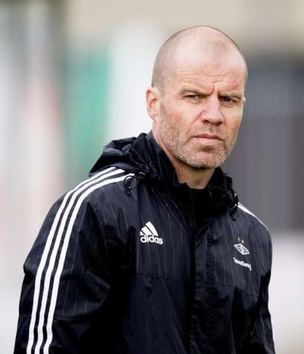 Ein kahlköpfiger Trainer in einer schwarzen Jacke steht auf einem Fußballfeld und trägt eine marineblaue Jacke mit weißen Streifen.