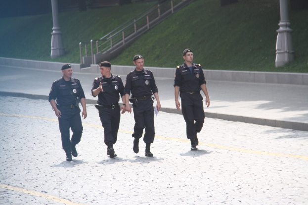 Drei Polizisten in Uniform und mit Mützen gehen auf einer Straße mit Pfählen, Geländern, Treppen und Gras im Hintergrund.