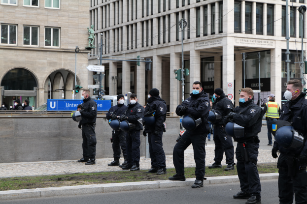 Eine Gruppe von Polizisten in schwarzen Uniformen und Masken steht vor einem Glasfenstergebäude mit Säulen in Berlin, Deutschland, einige halten Helme, mit Laternenmasten, Verkehrsampeln, Texttafeln, einer Statue, anderen Menschen und grasbewachsenem Boden im Hintergrund.