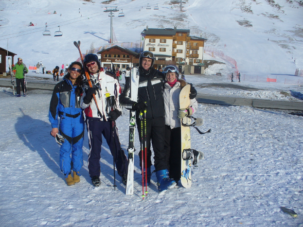 Vier Personen posieren für ein Foto mit Schlittschuhschutz, Werkzeugen und Gebäuden im Hintergrund vor einer schneebedeckten Berglandschaft.