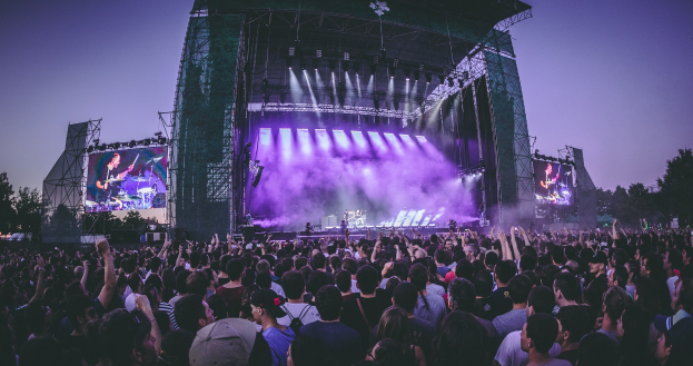 Eine große Menge Menschen bei einem Musikfestival schaut auf einer hell erleuchteten Bühne mit Instrumenten und Bildschirmen, die vor einem Gebäude, Bäumen und einem klaren blauen Himmel liegen.