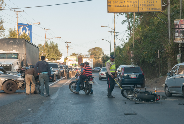Eine Gruppe von Menschen um ein verunglücktes Motorrad auf der Straße herumstehend mit mehreren Fahrzeugen, darunter ein Lastwagen, und einem Hintergrund aus Bäumen, Polen, Lampen, Brettern und Himmel.