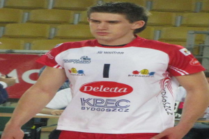 Ein Mann in einem roten und weißen Handballtrikot steht auf einem Feld, umgeben von Zuschauern, mit einem roten Banner im Hintergrund, auf dem "Krasnoyarsk vs Bydgoszcz" steht.