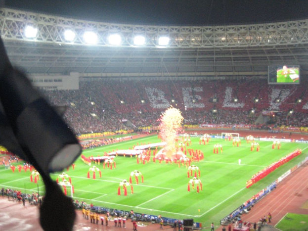 Ein Stadion mit Flutlicht, Eisengittern, Zuschauern, stehenden Personen, Werbetafeln, Anzeigetafeln, Ãberwachungskameras, Fahnen und Deko.