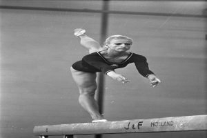 Eine Frau in einem Einteiler führt Gymnastik auf einer Stange aus, ihre Arme sind ausgestreckt und ihre Beine sind in einer anmutigen Kurve gebeugt, mit einem konzentrierten Gesichtsausdruck.