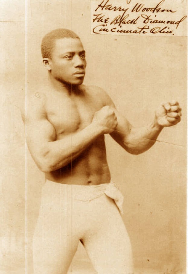 Ein altes Foto eines Mannes in einem Boxring, der Handschuhe und Shorts trägt und einen entschlossenen Gesichtsausdruck hat, kurze dunkle Haare und fokussierte Augen, aufrecht und stolz dasteht.