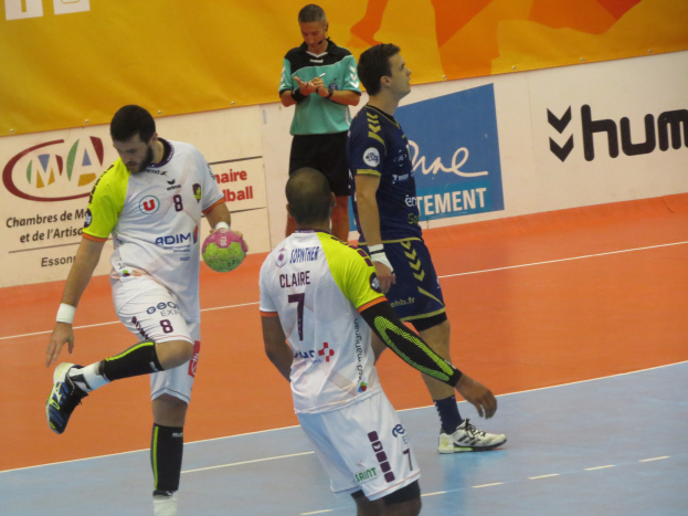 Eine Gruppe von Männern, die auf einem Handballfeld spielen, mit einem Mann, der einen Ball hält, und Bannern mit Text im Hintergrund.