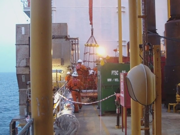 Ein Schiff mit M√§sten, Kisten, Gel√§ndern und Menschen mit Helmen, mit Wasser im Hintergrund.