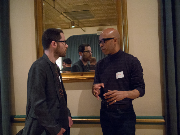 Ein afrikanischer Mann in schwarzem Hemd und Hose spricht mit einem Mann in schwarzem Mantel, mit einem Spiegel und brauner Wand im Hintergrund.