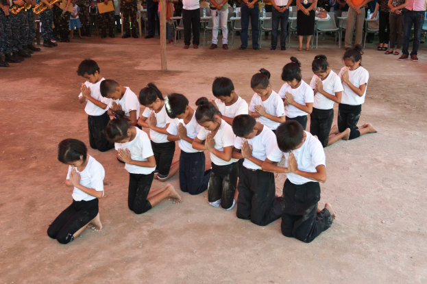 Eine Gruppe von Kindern kniet vor einer Menschenmenge, einige halten Musikinstrumente, mit Stühlen und einem Pfahl im Hintergrund, was auf eine Schulumgebung hindeutet.