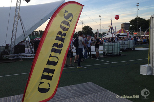 Lebendige Karnevalsszene mit Zelten, zentralem Mast, Menschen, Bäumen, Strommasten, Straßenbahnen und einem Eisenständer auf der linken Seite unter einem klaren Himmel.