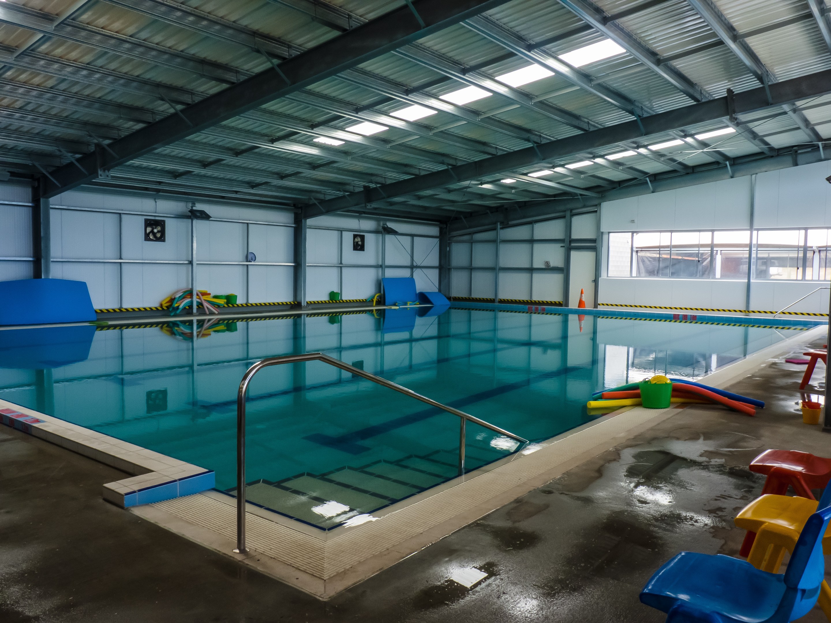 Indoor-Schwimmbad mit Stühlen an der rechten Seite, einem gefliesten Boden, einer Wand im Hintergrund und Deckenlampen.