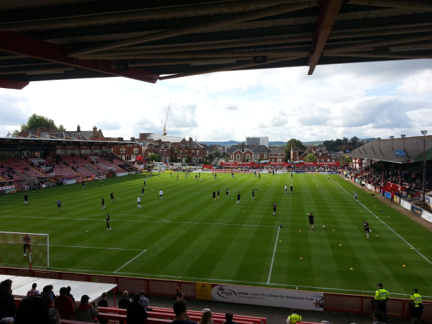 Ein Fußballspiel wird im St. Mary's Stadium gespielt, mit Zuschauern auf den Bänken und Stehplätzen, einem Torpfosten links und im Hintergrund Gebäude, Bäume und ein bewölkter Himmel.