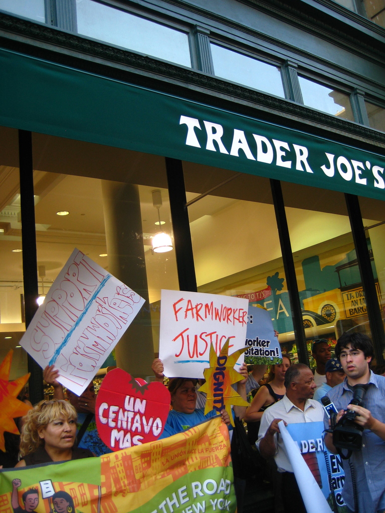 Eine Gruppe von Menschen steht in der Nähe eines Geschäfts und hält Schilder und Plakate in den Händen, wobei hinter ihnen eine Säule und Lampen auf dem Dach zu sehen sind.