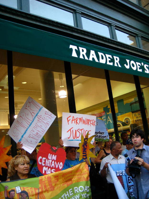 Eine Gruppe von Menschen steht in der Nähe eines Geschäfts und hält Schilder und Plakate in den Händen, wobei hinter ihnen eine Säule und Lampen auf dem Dach zu sehen sind.