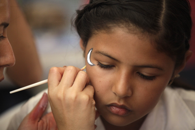 Ein Make-up-Artist trägt Make-up mit einem Pinsel auf das Gesicht eines jungen Mädchens auf, wobei beide Personen vor einem unscharfen Hintergrund im Fokus stehen.