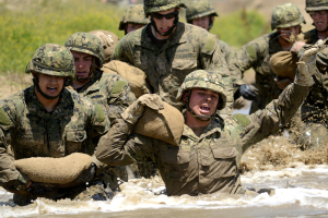 Eine Gruppe von Soldaten in Tarnkleidung rennt durchs Wasser, jeder trägt einen Sandsack, mit einem unscharfen Hintergrund, der schnelle Bewegung andeutet.