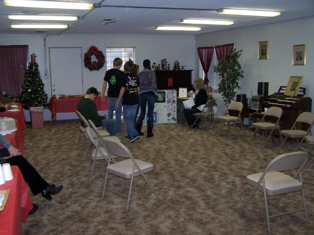Ein Raum mit mehreren Stühlen, Tischen und Menschen, einige stehen und einige sitzen. Auf den Tischen stehen Becher und Gefäße. Im Hintergrund befindet sich eine Wand mit einem Blumenstrauß, einem Vorhang, einer Tür, Fotorahmen und Fenstern. Ein Weihnachtsbaum und ein Piano sind ebenfalls vorhanden, mit Röhrenleuchten an der Decke.