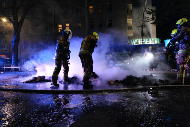 Feuerwehrleute in Helmen stehen nachts auf einer Straße neben einem qualmenden Rohr, mit Gebäuden, Bäumen und Fahrzeugen im Hintergrund und einer Tafel mit Schrift auf der rechten Seite.
