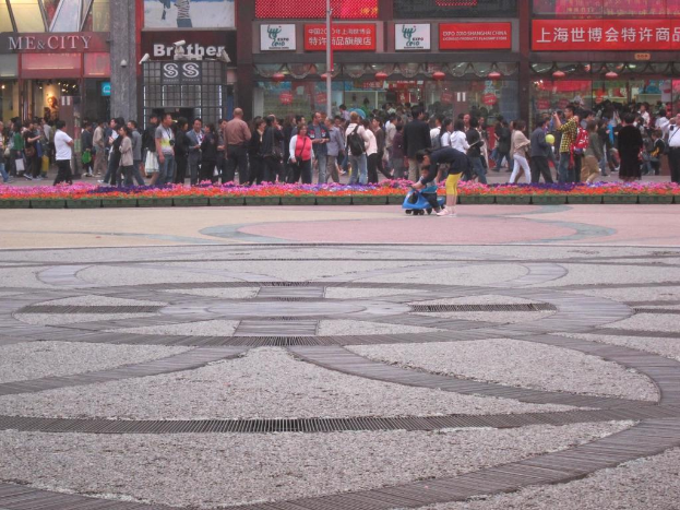 Ein Kind fährt auf einem Spielzeugfahrzeug im Vordergrund, während Menschen im Hintergrund und Schilder und Geschäfte im Hintergrund zu sehen sind.