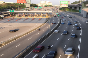 Eine vielbefahrene Autobahn mit zahlreichen Autos, Motorrädern, Verkehrskegeln, Schildern, Laternenmasten, einer Brücke, Bäumen, Gebäuden und einem klaren blauen Himmel.
