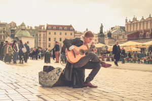 Ein Mann, der auf dem Boden sitzend eine Gitarre in einem belebten Stadtplatz spielt, umgeben von Füpfen und einer Tasche auf dem Weg, mit Gebäuden, einer Statue und einem klaren blauen Himmel im Hintergrund.