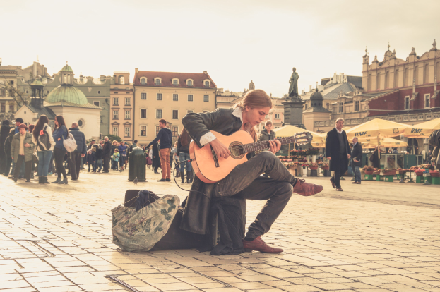 Ein Mann, der auf dem Boden sitzend eine Gitarre in einem belebten Stadtplatz spielt, umgeben von Füpfen und einer Tasche auf dem Weg, mit Gebäuden, einer Statue und einem klaren blauen Himmel im Hintergrund.