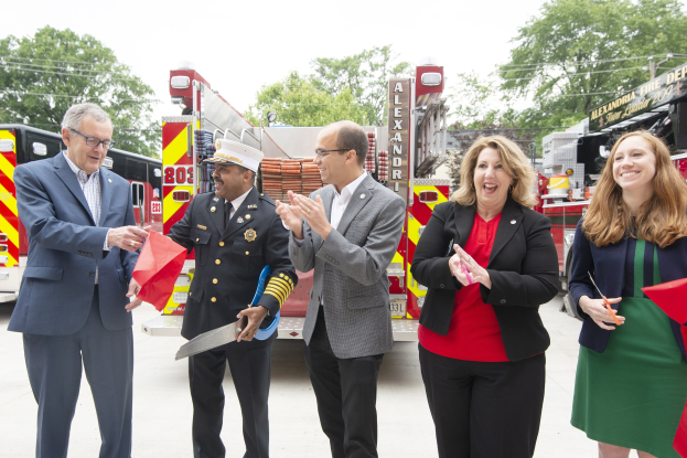 Gruppe von Menschen bei einer Ehrung für die Alexandria Fire Department, klatschend und lachend vor einem Feuerwehrauto mit Bäumen und einem klaren blauen Himmel im Hintergrund.