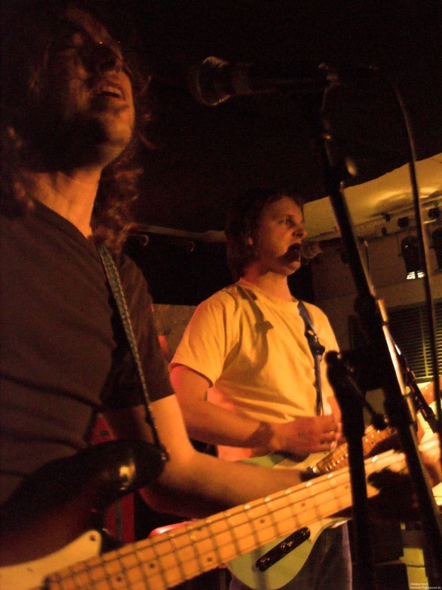 Eine Person in einem wei├čen Hemd singt in ein Mikrofon und spielt eine gr├╝ne Gitarre, neben einer anderen Person in einem schwarzen Hemd und Brille, die ebenfalls singt und eine schwarze Gitarre spielt, mit Scheinwerfern und anderen Instrumenten im Hintergrund.