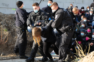 Gruppe maskierter Personen in Mützen, einige bewaffnet, die eine Frau umgeben, die von der Polizei begleitet wird, mit städtischer Vegetation und einem protestbezogenen Wandtext im Hintergrund.