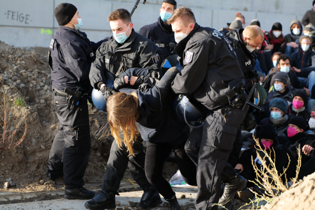 Gruppe maskierter Personen in Mützen, einige bewaffnet, die eine Frau umgeben, die von der Polizei begleitet wird, mit städtischer Vegetation und einem protestbezogenen Wandtext im Hintergrund.