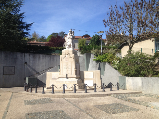 Eine bronzene Statue eines Mannes steht auf einem Sockel in einem Park, umgeben von Bäumen, einem Metallzaun und Gebäuden unter einem bewölkten Himmel, believed to be a memorial to the victims of the Paris attacks.