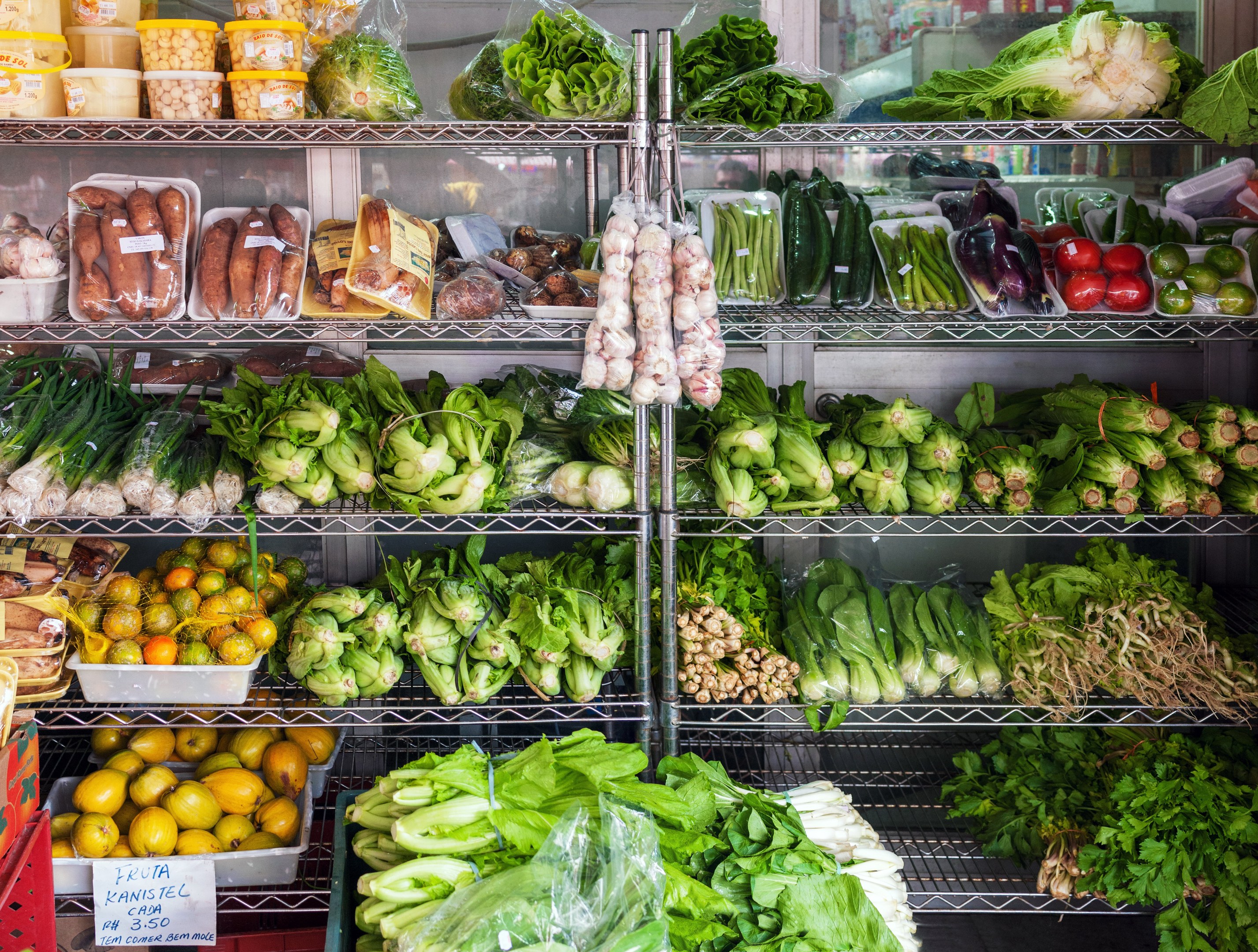Ein Gang in einem Supermarkt mit frischem Gemüse und Obst, verpackten Waren in Plastikhüllen, Kartons, einem Schild mit Text und einem Glasfenster im Hintergrund.