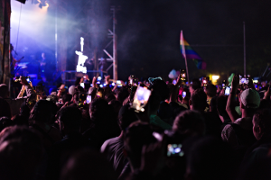Eine Menge bei einem Konzert hält beleuchtete Handys hoch, während eine kleine Band auf der Bühne unter hellen Lichtern spielt.