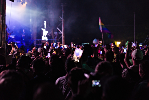 Eine Menge bei einem Konzert hält beleuchtete Handys hoch, während eine kleine Band auf der Bühne unter hellen Lichtern spielt.