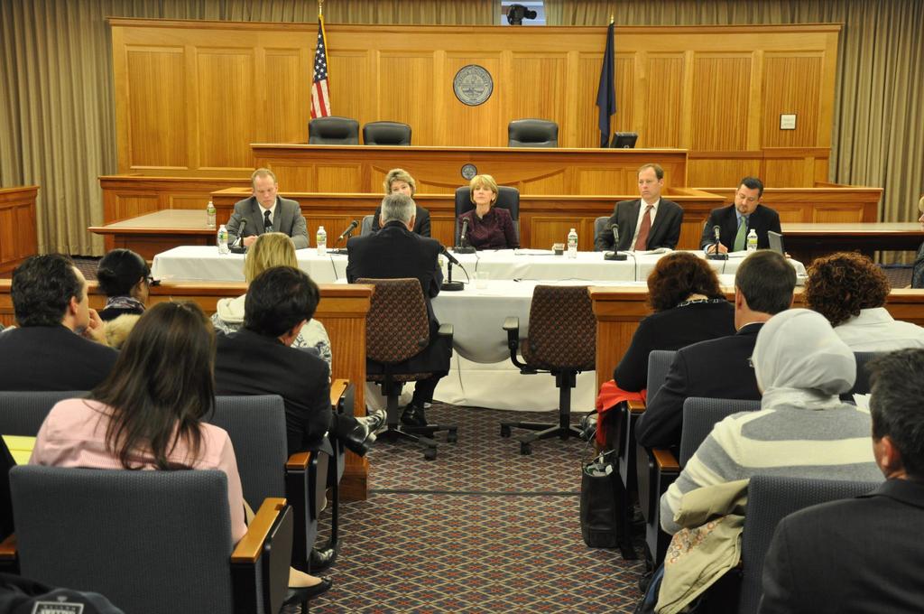 Ein Versammlungsraum mit einer Gruppe von Menschen, die auf Stühlen sitzen, zwei Flaggen anderer Länder, ein oranger Schreibtisch mit Wasserflaschen und einem Mikrofon.