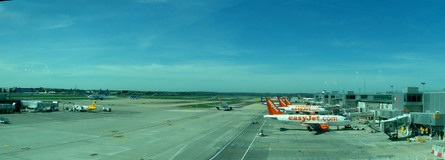 Eine Flughafen-Szene mit Flugzeugen, GebÃ€uden, Masten, Fahrzeugen und BÃ€umen, mit einem Himmel oben und einer StraÃe unten und einem Zaun im Vordergrund.
