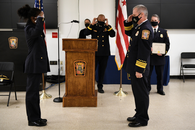 Gruppe uniformierter Personen in Masken auf einem Podium mit Mikrofon, umgeben von Fahnen und Möbeln, während einer Vereidungszeremonie für einen neuen Feuerwehrchef.