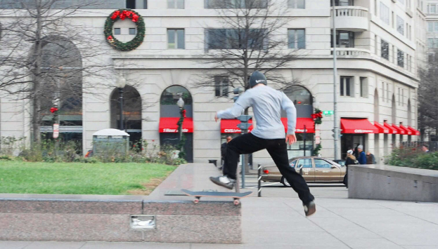 Ein Mann springt von einem Skateboard auf eine grasbewachsene Fläche, mit Menschen auf einer nahen Straße und einem geparkten Auto, mit Bäumen und Gebäuden im Hintergrund.