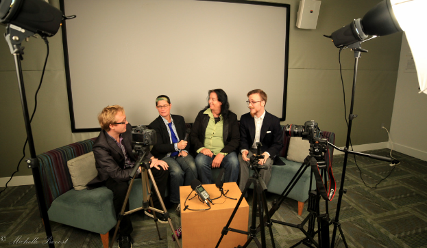 Eine Gruppe von drei Personen sitzt auf einem Sofa und gibt ein Interview. Links befindet sich eine Person in einem schwarzen Mantel und Hose, in der Mitte eine Person in einem Mantel und einer blauen Krawatte, die ein Mikrofon hält, und rechts eine strahlende Person. Auf dem Tisch in der Mitte befindet sich ein Mikrofonrekorder, Kameras sind auf der rechten und linken Seite und große Scheinwerferständer sind vorhanden, sowie eine Projektionswand im Hintergrund.