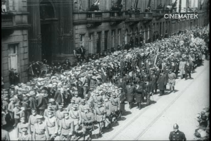 Schwarzes Foto einer Parade mit einer großen Menge Menschen, die vor einem Gebäude die Straße entlanggehen, einige halten Gewehre.