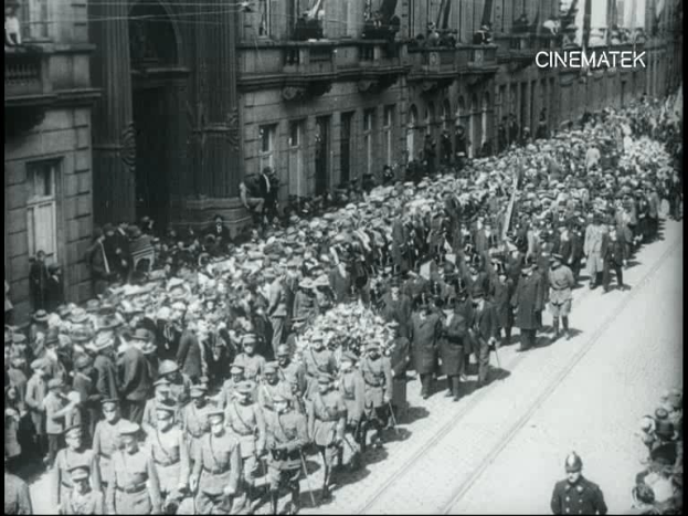 Schwarzes Foto einer Parade mit einer großen Menge Menschen, die vor einem Gebäude die Straße entlanggehen, einige halten Gewehre.