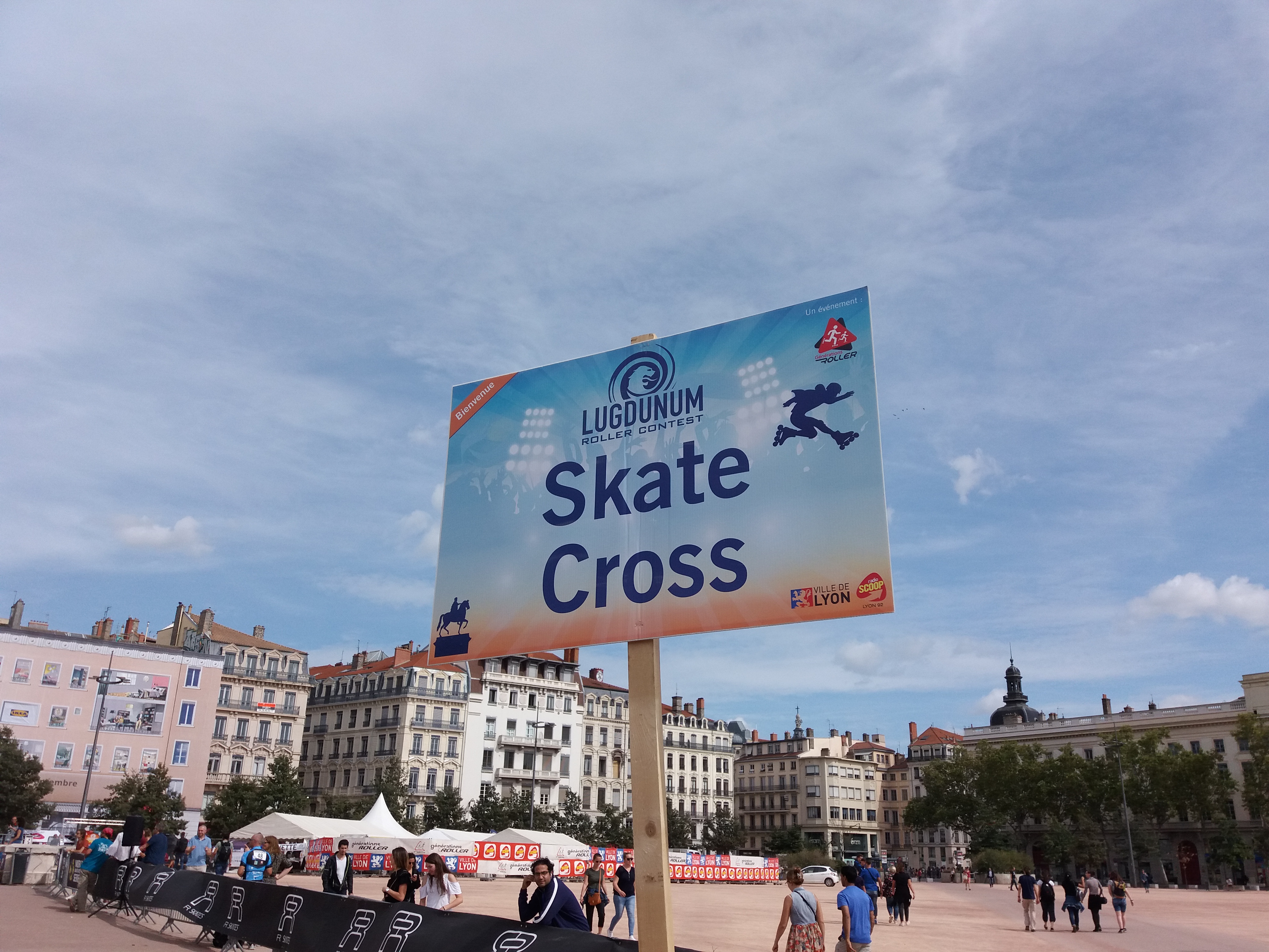 Ein großes Plakat in einem Stadtplatz umgeben von Menschen, Bannern, Zelten, einem Zaun, Straßenlaternen, Bäumen und Gebäuden unter einem bewölkten Himmel.