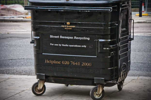 Ein schwarzer Mülltonnenbehälter steht in der Mitte des Bildes mit einer Straße unten.