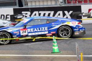 Ein blau-weißer Rennwagen fährt auf einer Rennstrecke, mit einem Verkehrskegel im Vordergrund und Schildern mit Text, Geländern und anderen Gegenständen im Hintergrund.