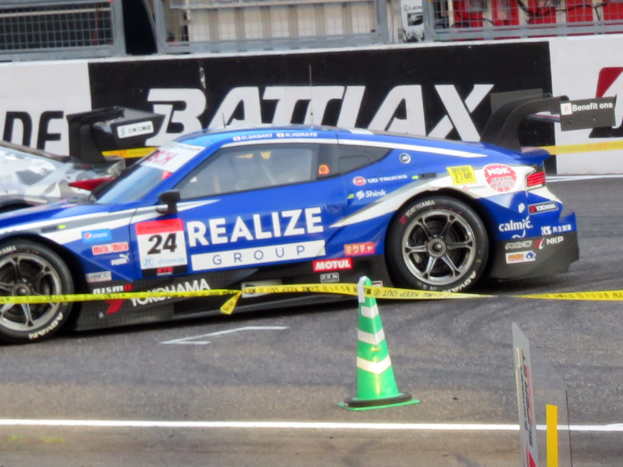 Ein blau-weißer Rennwagen fährt auf einer Rennstrecke, mit einem Verkehrskegel im Vordergrund und Schildern mit Text, Geländern und anderen Gegenständen im Hintergrund.